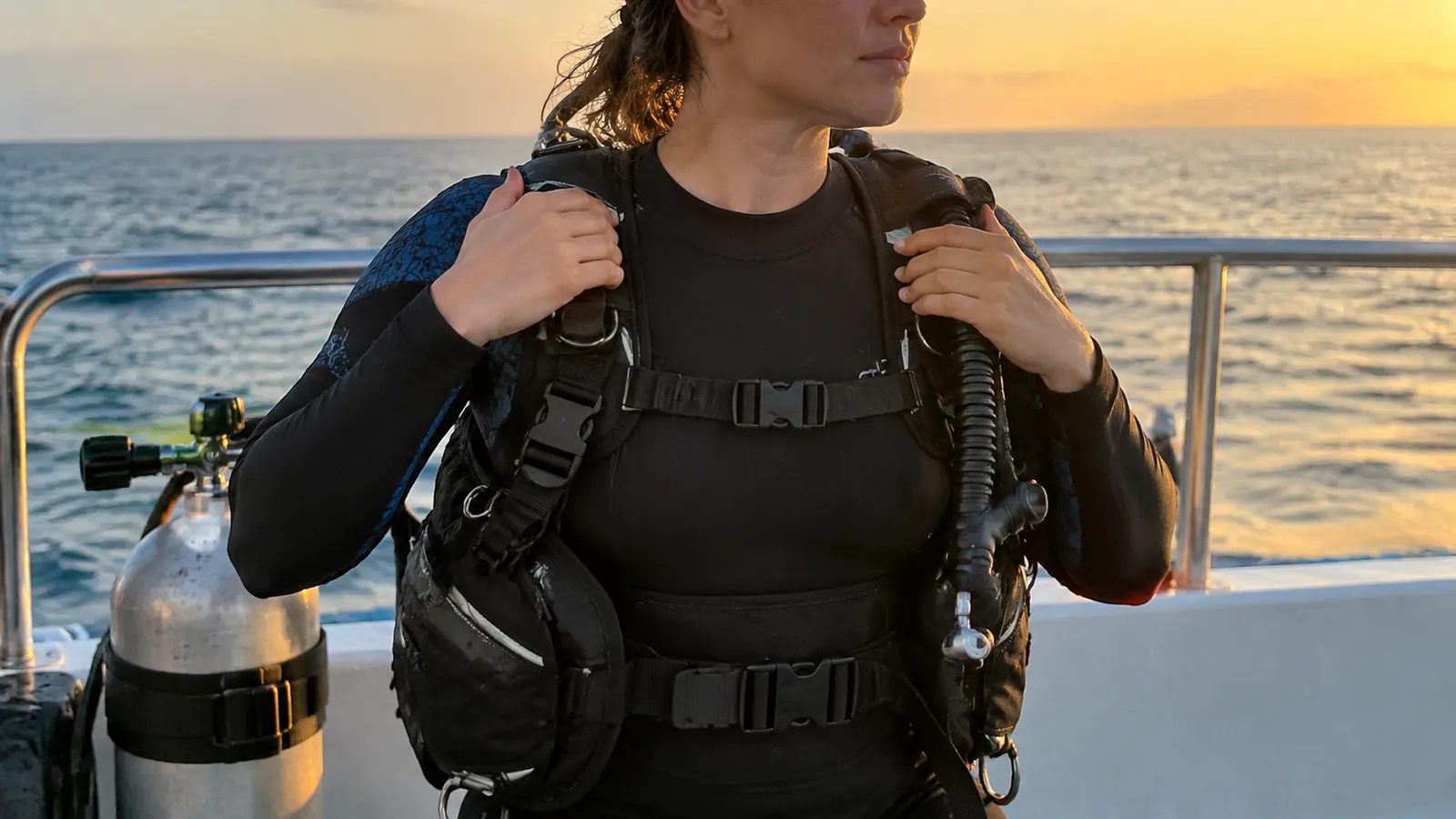 Diver on a boat deck pulling the shoulder straps of a back-inflation BCD snug over a black rash guard, tank standing beside them against the rail