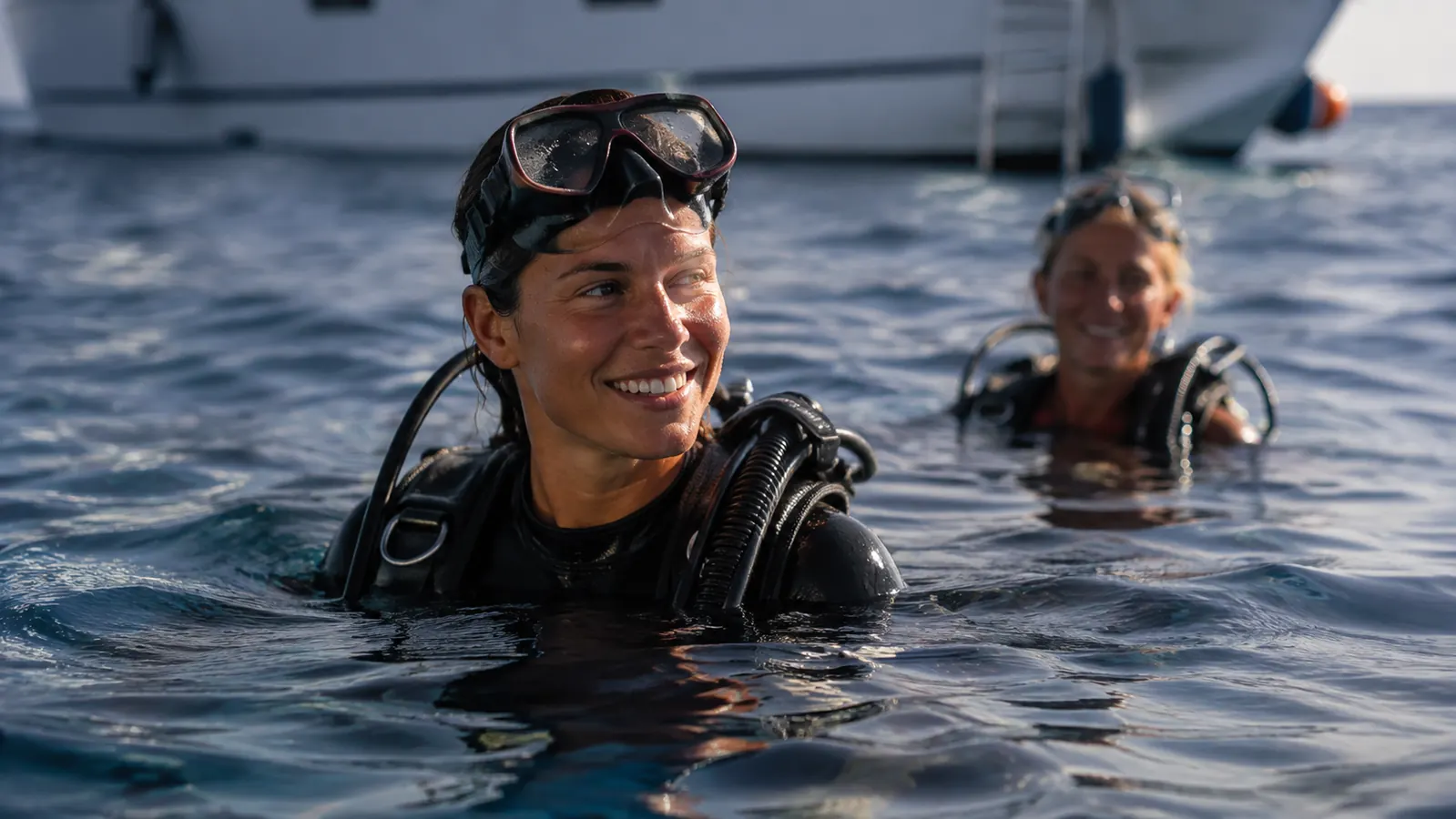 Two divers resting at the surface between dives, one floating upright and relaxed in a jacket-style BCD with the mask pushed up on the forehead, calm blue sea and a dive boat behind, soft late-afternoon light