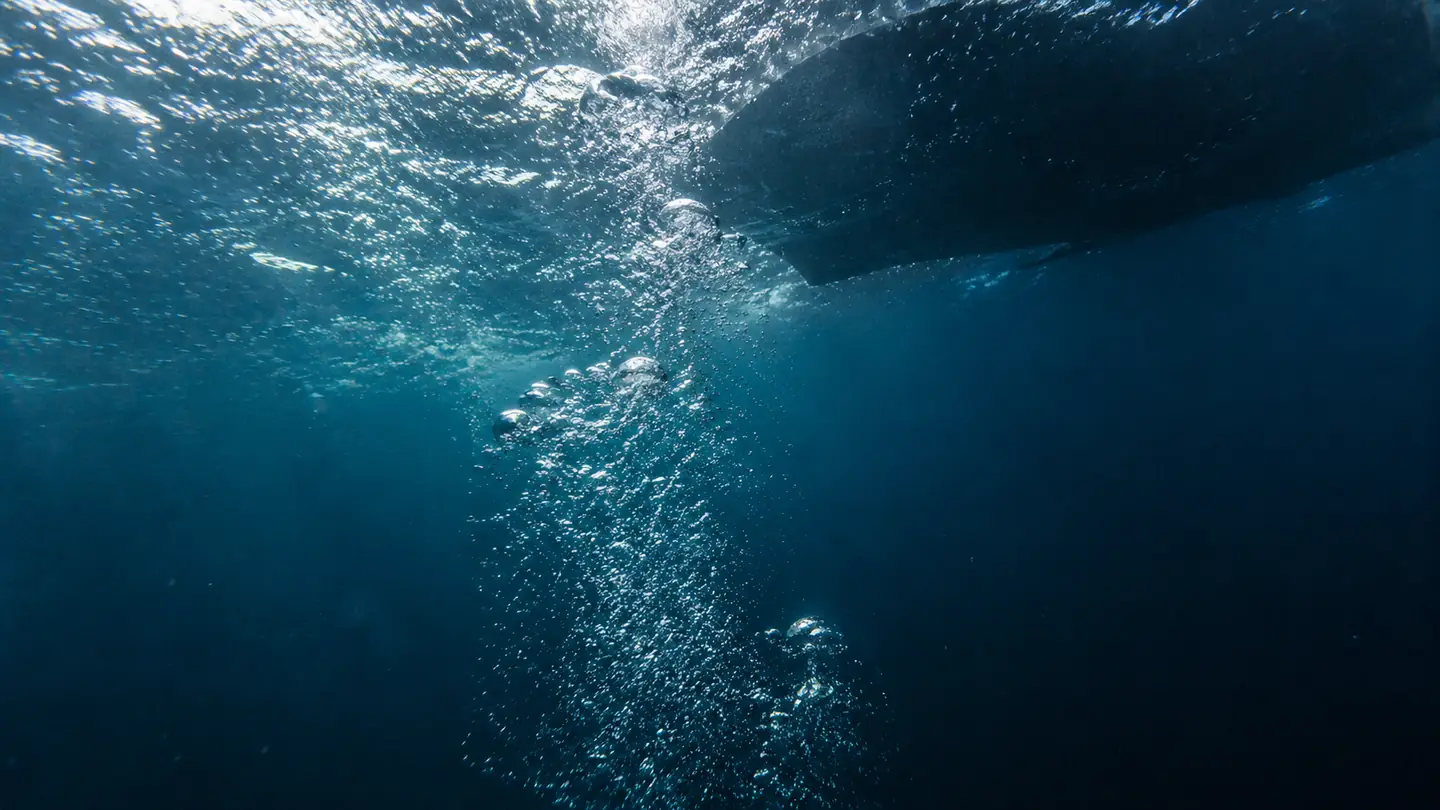Looking up from a safety stop, bubbles rising toward a bright sunlit surface with the boat hull shadowed above