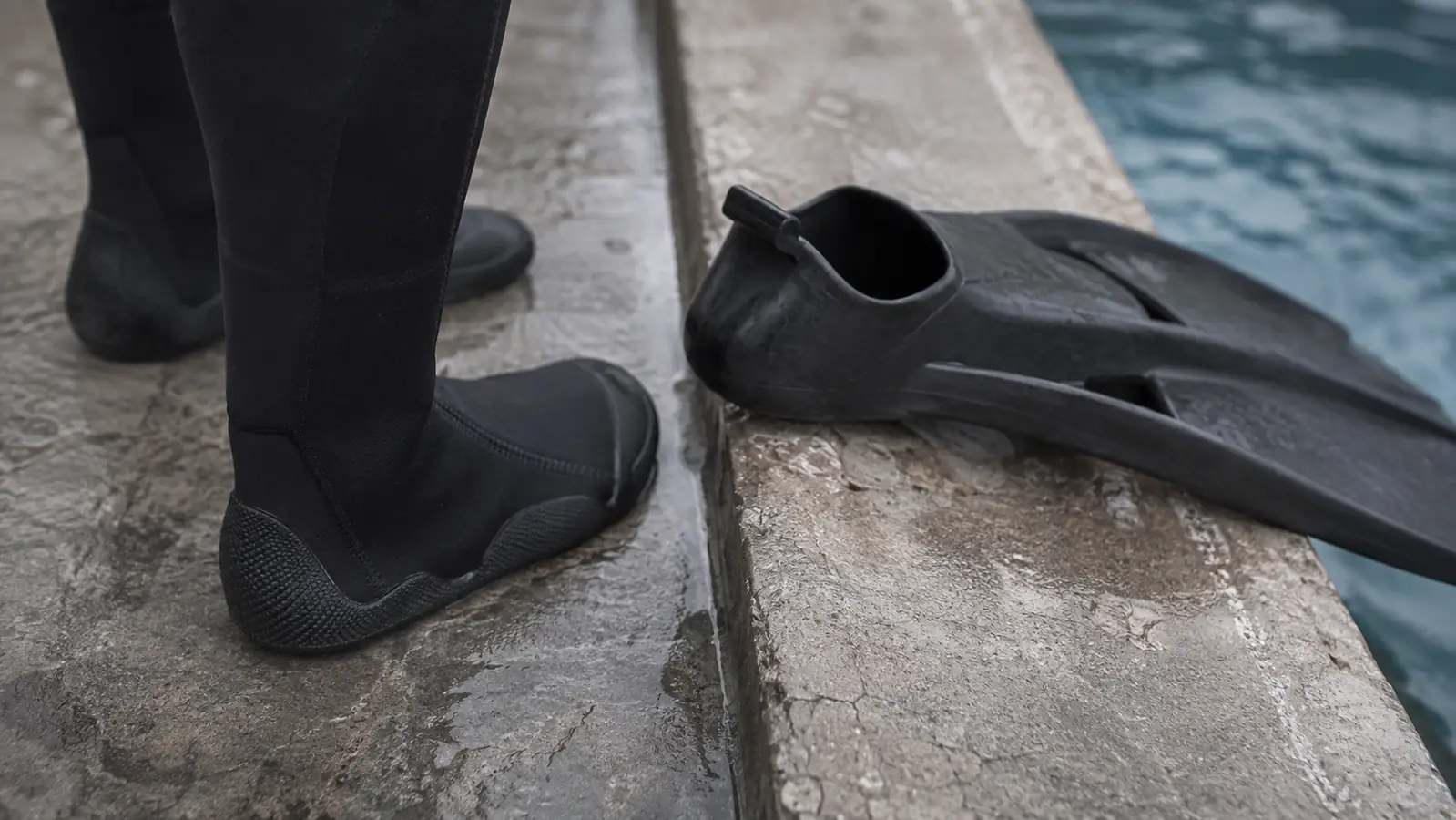 Feet in thick black neoprene boots standing on a wet concrete pool deck, a single open-heel fin resting on the pool coping next to clear water