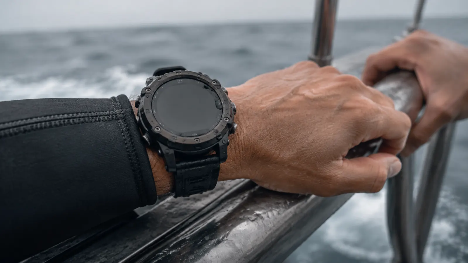 Close-up of a dive computer on a diver's wrist, forearm resting on a boat rail with salt spray on the casing, screen angled toward the camera.