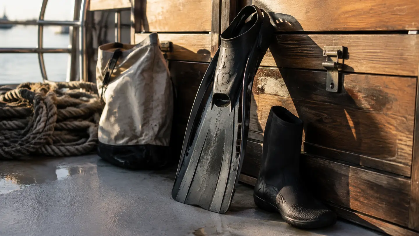 A scuba fin and a neoprene boot propped against a weathered wooden dive locker on a boat deck in late afternoon light